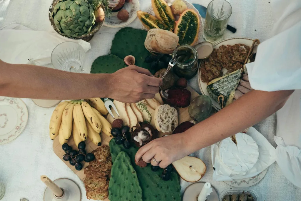 Hands reach over a colorful spread of fresh fruits and vegetables on a picnic table.